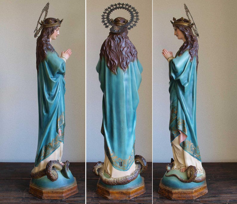 K&ouml;nnte beinhalten: Vintage Statue einer Frau in einem flie&szlig;enden tealfarbenen Gewand, geschm&uuml;ckt mit einer goldenen Krone und Heiligenschein. Die Statue steht auf einem Sockel mit einer sich windenden Schlange. Die H&auml;nde der Figur sind zum Gebet gefaltet.