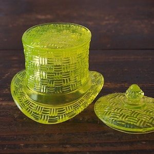 May include: A yellow-green glass top hat-shaped container with a matching lid. The container and lid have a woven basket-like texture. The lid is oval-shaped with a small knob on top. The container sits on a dark wooden surface.