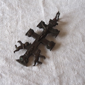 May include: A dark brown metal double-barred cross pendant featuring a central crucifix figure and two small cherub figures positioned on the lower crossbar. The cross has a small metal loop at the top for attachment to a chain or cord.