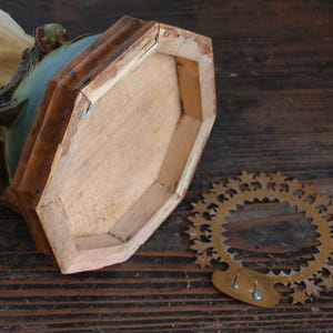 K&ouml;nnte beinhalten: Detaillierte Ansicht eines Vintage-Sockels aus Holz, der die Unterseite mit einer hellbraunen Oberfl&auml;che und einem dekorativen Metallring zeigt. Die Skulptur hat ein t&uuml;rkis-beige Farbschema mit sichtbaren Alters- und Gebrauchsspuren.