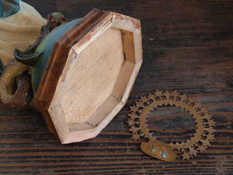 K&ouml;nnte beinhalten: Detaillierte Ansicht eines Vintage-Sockels aus Holz, der die Unterseite mit einer hellbraunen Oberfl&auml;che und einem dekorativen Metallring zeigt. Die Skulptur hat ein t&uuml;rkis-beige Farbschema mit sichtbaren Alters- und Gebrauchsspuren.