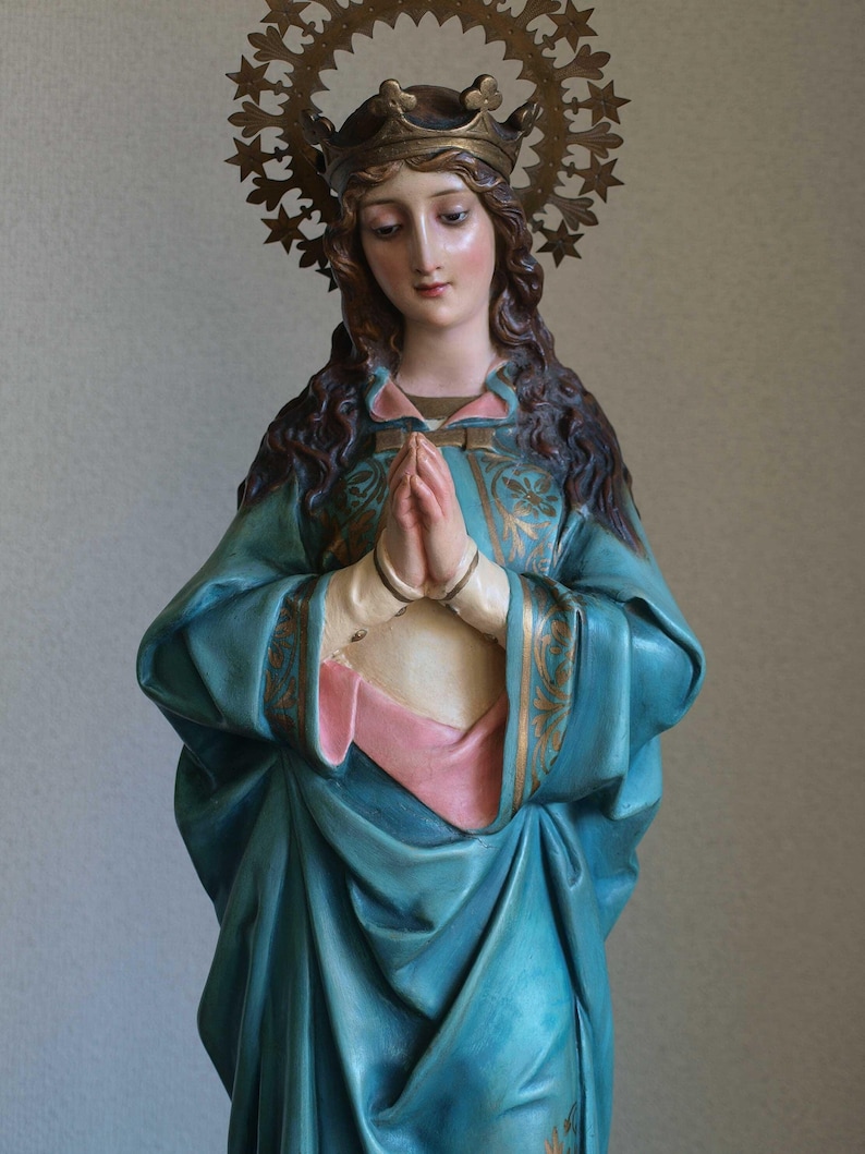 K&ouml;nnte beinhalten: Statue einer Frau mit Krone und Heiligenschein, die ein blaues Gewand mit goldenen Akzenten und ein rosa Untergewand tr&auml;gt. Ihre H&auml;nde sind zum Gebet gefaltet. Die Statue hat einen friedlichen Gesichtsausdruck.