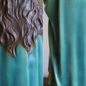 K&ouml;nnte beinhalten: Detailansicht einer Statue, die den R&uuml;cken von Kopf und Schultern zeigt. Die Statue hat langes, welliges braunes Haar und ein teal-farbenes Gewand. Der Stoff weist subtile Farb- und Texturvariationen auf.