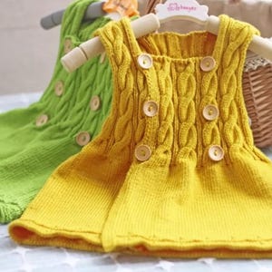 Può includere: Due vestitini di maglia per neonati, uno verde e uno giallo, con bottoni di legno e un motivo a treccia. I vestitini sono appesi a un appendiabiti di legno.