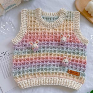 May include: A rainbow striped crocheted vest with a waffle stitch pattern and three small crocheted bunny appliques. The vest has a white trim and a small tag that says "Story Small Town Store".
