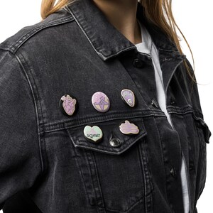 May include: A black denim jacket adorned with six enamel pins. The pins feature various designs, including a heart, a lightning bolt, and the word "OUTER". The pins are in shades of pink, purple, and green.