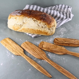 May include: A set of four wooden spatulas with distinct grain patterns arranged near a loaf of bread resting on a grey and white striped cloth. The spatulas feature long, flat handles and wide, rectangular heads with rounded corners and edges.