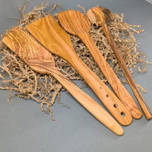 May include: A set of three wooden kitchen utensils featuring distinct grain patterns. The collection includes a wide flat spatula with three circular holes in the handle, a rounded spatula, and a long slender spoon with a circular bowl end.