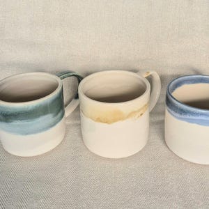 May include: Three ceramic mugs with a cylindrical shape and a handle on the side. The left mug features a green and blue gradient glaze, the centre mug has a tan and cream glaze, and the right mug displays a blue rim over a cream base.