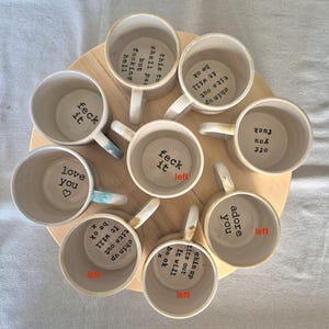 May include: A collection of eight ceramic mugs arranged in a circle, each featuring black stamped text on the interior base. Phrases include 'feck it', 'love you', 'this too shall pass but fucking hell', 'chin up tits out it will be ok', and 'adore you'.