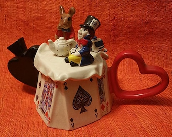Könnte beinhalten: Eine Teekanne aus Keramik in Form eines Spielkartenhauses mit einem roten Herzgriff. Die Teekanne zeigt ein weißes Kaninchen, eine Figur des Hutmachers und eine kleine Teekanne oben drauf. Die Teekanne ist mit Spielkarten verziert.