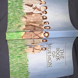May include: A hand-painted book cover featuring a group of people in brown robes standing in a field of green grass. The title "THE BOOK OF MORMON" is written in gold lettering on a blue background.