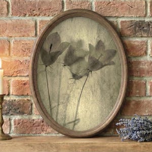 May include: An oval framed botanical print featuring sepia-toned floral silhouettes. A lit candle in a brass holder and a bundle of lavender sit on a wooden surface against a brick wall.
