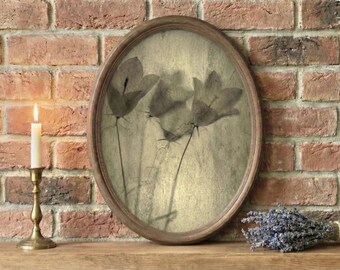 Könnte beinhalten: Ein oval gerahmter botanischer Druck mit sepiafarbenen Blumensilhouetten. Eine brennende Kerze in einem Messinghalter und ein Lavendelstrauß stehen auf einer Holzoberfläche vor einer Backsteinmauer.