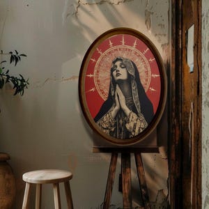 Könnte beinhalten: Ein oval gerahmter Druck einer Frau mit gefalteten Händen im Gebet. Das Kunstwerk zeigt einen roten Hintergrund mit einem goldenen Heiligenschein-Design. Die Frau trägt einen schwarzen Schleier und ein gold gemustertes Gewand. Der Rahmen ist dunkelbraun.