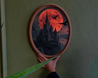 Puede incluir: Una impresión ovalada enmarcada en madera representa un castillo gótico silueteado contra una gran luna roja. Pájaros negros vuelan en el cielo. La impresión se sostiene contra una pared verde.