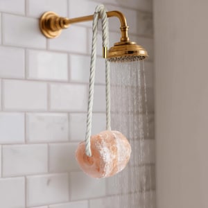 Op de afbeelding: Een messing douchekop met stromend water en een roze Himalaya zoutsteen hangend aan een wit touw. De douchekop is bevestigd aan een witte betegelde muur. De zoutsteen heeft een natuurlijke, getextureerde vorm.