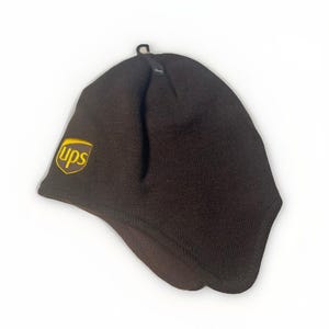 May include: A brown knit hat with ear flaps. The hat features the UPS logo in gold on the side. The hat is designed to keep the wearer warm and protected from the elements.