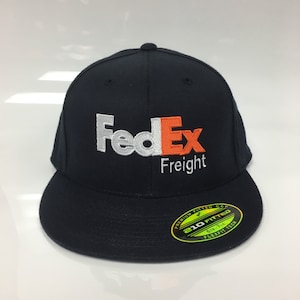 May include: A navy blue fitted baseball cap with a white and orange FedEx Freight logo embroidered on the front. The cap has a green Flexfit label on the side.