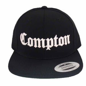 May include: Black baseball cap with a white embroidered "Compton" in old English lettering. The cap has a black adjustable strap with a white "The Classics Snapback" label.
