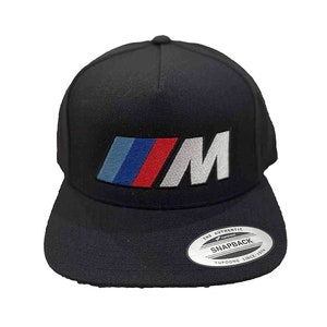 May include: Black and white embroidered "M" logo on a black baseball cap with a blue, red, and white stripe design. The cap has a black bill and a snap closure. The label on the back reads "The Authentic Yupoong Snapback Since 1974."