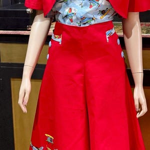 May include: A retro outfit comprising a red skirt and bolero jacket. The outfit includes a light blue collared shirt with a colourful aeroplane pattern. The red skirt has a matching aeroplane design and blue button details.