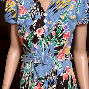 May include: A light blue vintage dress with a colourful floral pattern, including red, pink, yellow, and purple flowers, set against black and green foliage. The dress has a V-neck, short sleeves, and a matching belt. White buttons fasten the bodice.