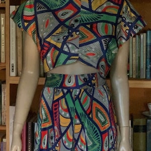 Può includere: Un completo a due pezzi composto da un top corto a maniche corte e pantaloni a vita alta, entrambi caratterizzati da un vivace motivo geometrico nei toni del verde, rosso, giallo e blu, con contorni blu scuro su un tessuto plissettato.