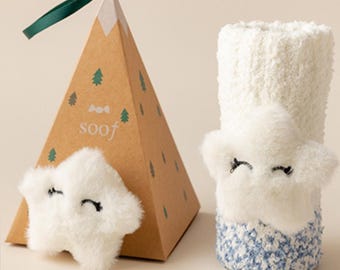 May include: A pair of white fuzzy socks with a star-shaped face design and blue accents. A brown triangular gift box with the word "soof" and green tree illustrations is next to the socks. A green ribbon is attached to the top of the box.