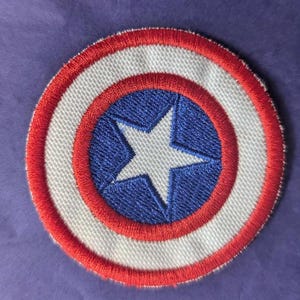 Può includere: Un cerotto ricamato in rosso, bianco e blu con una stella bianca su un cerchio blu circondato da un cerchio bianco e un cerchio rosso esterno.