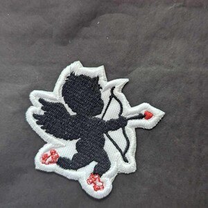 May include: Embroidered patch featuring a black Cupid figure with a bow and arrow, set against a white background with a decorative edge. The Cupid's feet and arrow tip are red. Ideal for clothing or accessories.