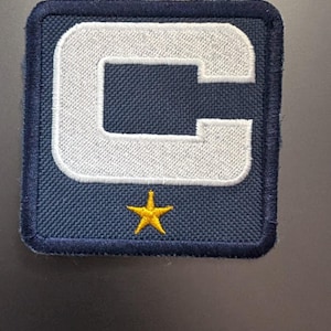 Könnte beinhalten: Ein marineblauer Patch mit einem weißen Buchstaben "C" und einem gelben Stern in der Mitte.