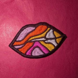 Könnte beinhalten: Ein farbenfroher gestickter Patch in Form einer Lippe. Die Lippen sind schwarz umrandet und mit einem wirbelnden Muster aus Orange, Gelb, Rosa und Lila gefüllt.