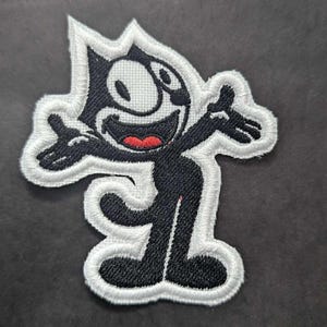 Könnte beinhalten: Ein gestickter Aufnäher von Felix the Cat, einer Zeichentrickfigur mit einem breiten Grinsen und ausgestreckten Armen. Der Aufnäher ist schwarzweiß mit einem weißen Rand und befindet sich auf einem dunkelgrauen Hintergrund.