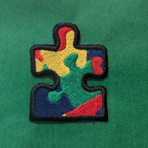 May include: A colourful puzzle piece patch with red, yellow, green, and blue sections. The patch is embroidered with black thread and has a black outline.
