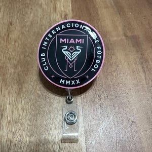 May include: A black and pink round badge with the text "Club Internacional de F&uacute;tbol Miami MMXX" and a pink flamingo logo.