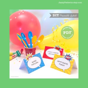 May include: DIY printable labels with comic book theme. The labels have text like "Super Popcorn", "X-Ray Vision", and "Kryptonite Punch". Colorful balloons and spoons are in the background. The labels are designed for parties.