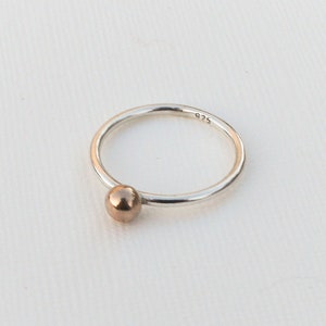 May include: A minimalist silver ring with a small, rose gold-coloured sphere accent. The ring has a thin band and a simple, elegant design. The ring is likely made of sterling silver, as indicated by the "925" stamp.
