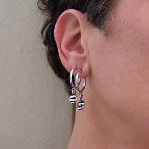 Puede incluir: Un par de pendientes de aro de plata con pequeños dijes circulares texturizados que cuelgan de la parte inferior de cada aro. Los pendientes están pulidos y tienen una superficie reflectante, con dijes que muestran un patrón ondulado.