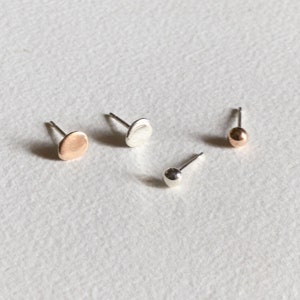 May include: Four small stud earrings arranged on a textured surface. Two earrings feature flat, circular discs, one in a rose gold tone and one in silver. The other two earrings are spherical ball studs, one in a rose gold tone and one in silver.