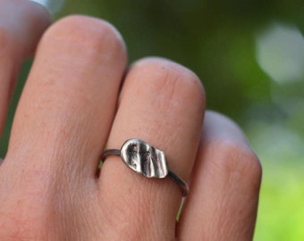 May include: A thin, dark-toned metal ring featuring a small, irregular, textured focal piece with a rippled surface. The metal has a matte, oxidized finish, and the central element is shaped like a slightly elongated, organic, flattened pebble.