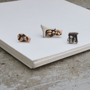 May include: Three pieces of jewelry are displayed on a white surface. One is a small, textured, bronze-colored stud. Another is a silver and bronze-colored pendant. The third is a silver stud with a bronze-colored design. The jewelry is set against a neutral background.
