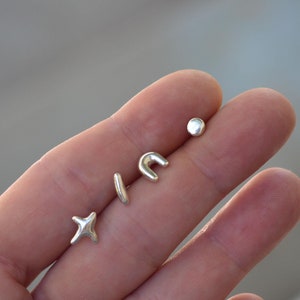 May include: A set of four small silver stud earrings with smooth, rounded surfaces. The shapes include a four-pointed star, a thin vertical bar, a curved arch, and a small circular dot, all featuring a polished metallic finish and simple design.