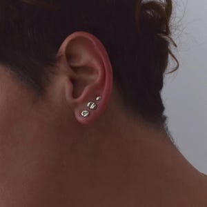 May include: Three silver-toned stud earrings arranged in a vertical line on an earlobe. The earrings are circular and feature a flat, polished surface with a subtle, indented line detail across the center of each piece, resembling a screw head.