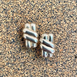 May include: A pair of silver-toned, geometric clip-on earrings. Each earring features a stacked, curved design with a polished finish. The earrings are displayed on a textured, cork-like surface.