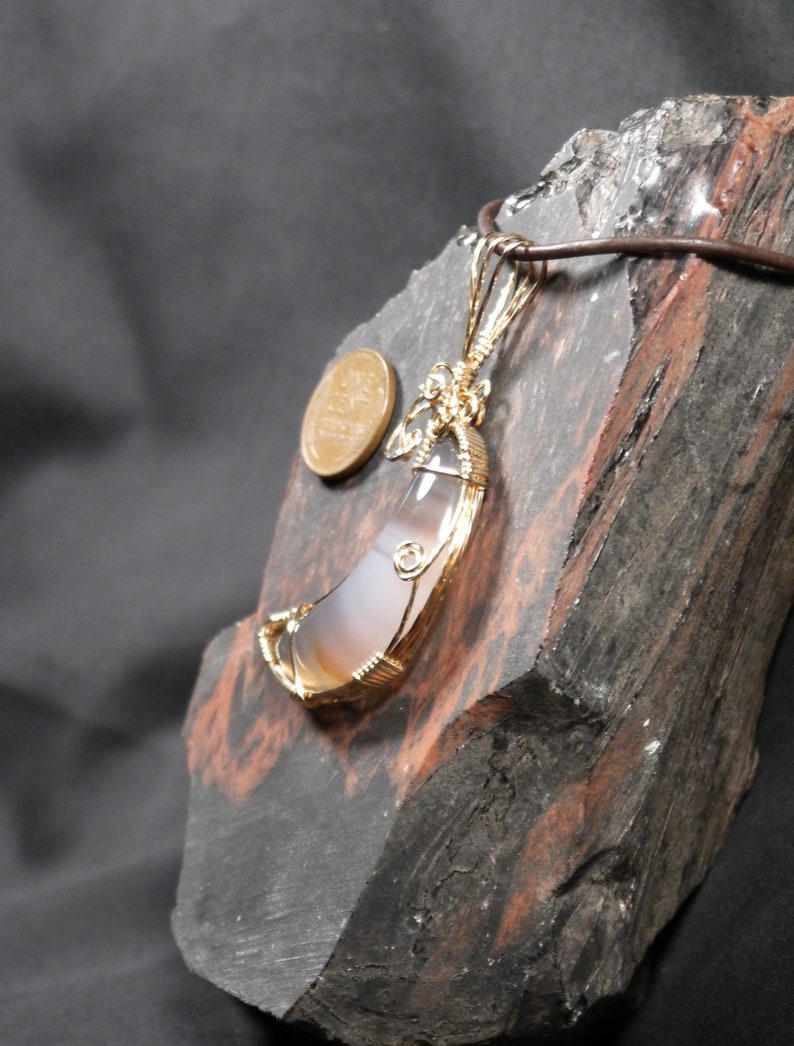 K&ouml;nnte beinhalten: Ein Anh&auml;nger aus Achat in Halbmondform, umwickelt mit goldfarbenem Draht. Der Anh&auml;nger wird auf einem dunkelbraunen St&uuml;ck Holz pr&auml;sentiert.