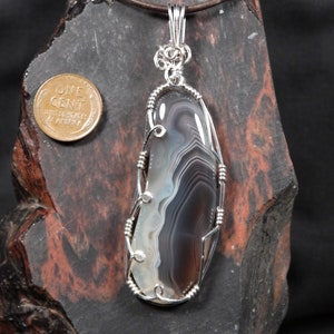 May include: A silver wire wrapped pendant featuring a gray and white agate stone. The pendant is hanging from a brown leather cord and is displayed on a dark brown rock.