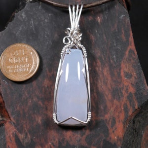 May include: A handcrafted pendant featuring a pale blue gemstone encased in a silver wire wrap. The pendant is suspended from a brown leather cord. A US one-cent coin is next to the pendant for size comparison.