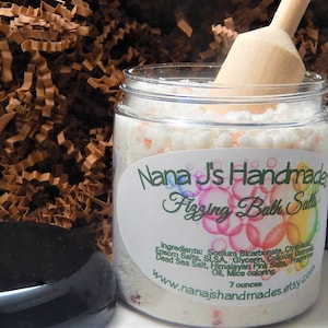 May include: A clear jar filled with white and pink bath salts, with a wooden scoop inside. The jar has a label that reads "Nana J's Handmade Fizzing Bath Salts." The jar contains 7 ounces of bath salts.