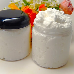 May include: Two clear jars filled with white, fluffy product, one with a black lid and the other open. The jars are in front of a colorful floral arrangement with orange, yellow, and pink flowers. The product appears to be a cosmetic item.
